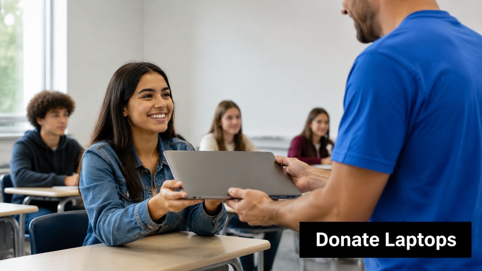 A student smiling while receiving a donated laptop from a man in a blue t-shirt in class.