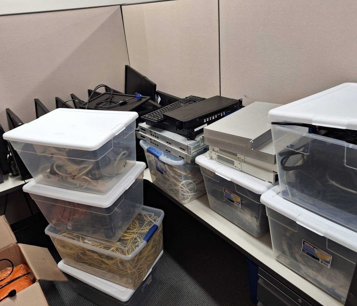Stacks of clear plastic bins filled with network cables and electronic equipment await E-waste disposal in an office corner. A laptop and additional devices rest on top, surrounded by a few cardboard boxes and power cords, all part of the electronics recycling process.