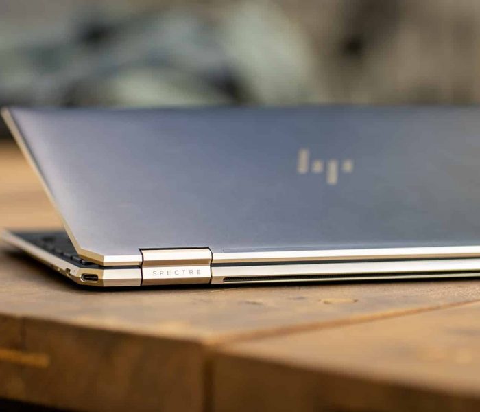 A sleek, partially open HP Spectre laptop sits on a wooden table. The laptop has a metallic finish and a reflective logo on the lid. The hinge design suggests it is a convertible model that can be folded into different modes. When upgrading, consider electronics waste disposal to recycle laptops responsibly.