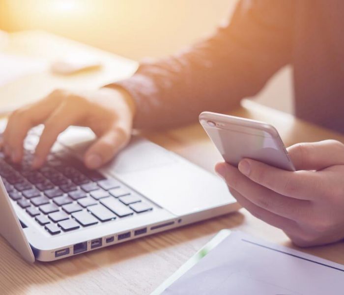 A person wearing a dark shirt is seated at a desk, typing on a laptop with their left hand while holding a smartphone in their right hand. Sunlight softly illuminates the scene, where responsible electronics recycling can help reduce e-waste. A document lays partially visible on the desk.